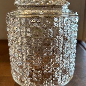 May include: A clear glass jar with a textured, patterned surface. The jar has a lid and a rounded shape. The surface is covered in a repeating pattern of squares and circles. The jar is likely used for storage or display.