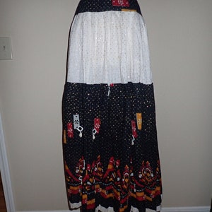 May include: A long, tiered skirt with a white, black and gold polka dot pattern. The skirt has a black waistband and a red, yellow and black patterned trim at the bottom.
