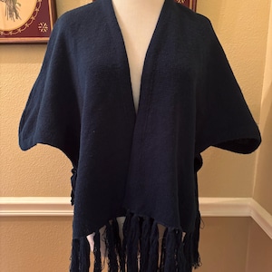 May include: A navy blue shawl with a draped design and fringe detailing. The shawl is displayed on a mannequin, showcasing its open front and textured fabric. The fringe hangs along the bottom edge.