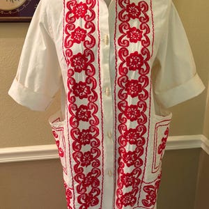 May include: White, short-sleeved tunic with red floral embroidery. The tunic features a mandarin collar, button-down front, and two pockets. The red embroidery is a prominent design element, creating a striking contrast against the white fabric.