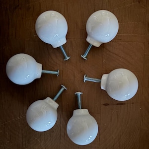 May include: Six white ceramic cabinet knobs with a glossy finish, arranged in a circular pattern. Each knob has a rounded shape and a silver-colored screw for attachment. The knobs are set against a warm, brown wooden surface.