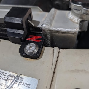 May include: A black plastic radiator shroud bracket featuring a red Z logo and a silver metal bolt, mounted on a beige automotive panel next to a metallic radiator with a cap. A silver sticker with black text is visible on the beige panel.