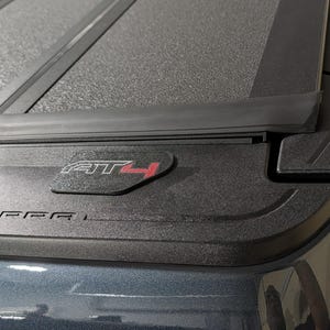May include: Close-up of a black truck bed cover with the text "AT4" in red and "SIERRA" in silver. The cover has a textured surface and a sleek, modern design.