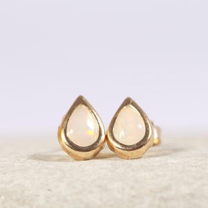 Op de afbeelding: Een paar druppelvormige oorstekers. Elke oorbel heeft een parelmoer, crèmekleurige steen gezet in een goudkleurige bezel. De oorbellen worden gepresenteerd op een neutraal, getextureerd oppervlak met een zachte, onscherpe achtergrond.
