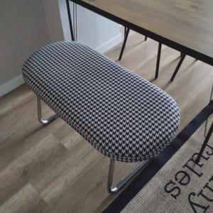 May include: An upholstered bench with a black and white houndstooth pattern. The bench has a curved, oval shape and sits on chrome legs. A wooden table with black metal legs is in the background.