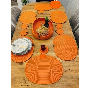 May include: An orange table setting featuring oval placemats, coasters, and a central bowl. The set includes plates, silverware, and a small glass. The woven texture and pumpkin-shaped accents create a cohesive look.