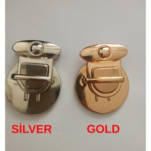 May include: Two metal purse clasps, one silver and one gold, are displayed against a white background. The clasps have a rounded shape with a central bar and a decorative top. The word "SILVER" is written in red below the silver clasp, and "GOLD" below the gold clasp.