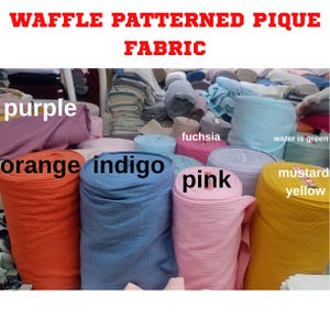 Può includere: Una varietà di rotoli di tessuto piqué a motivo waffle colorati. I colori includono arancione, indaco, rosa e giallo senape. L'immagine mostra anche rotoli di tessuto viola, fucsia e azzurro. Il testo in alto recita "WAFFLE PATTERNED PIQUE FABRIC."