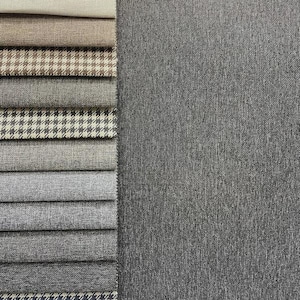 May include: A collection of fabric swatches in various shades of gray, beige, and brown, with houndstooth and solid patterns. The swatches are stacked next to a larger piece of gray textured fabric, showcasing different textures and designs for upholstery or textile projects.