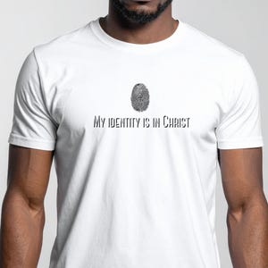 Könnte beinhalten: Weißes T-Shirt mit dem Text "My Identity Is In Christ" in schwarzer, fetter Schrift. Oberhalb des Textes befindet sich ein Fingerabdruck-Design mit einem Kreuz in der Mitte. Das Shirt hat kurze Ärmel und scheint aus Baumwolle zu sein.