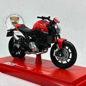 May include: A red and black Ducati Monster motorcycle scale model featuring a red fuel tank with the text Monster, a black frame, black wheels, and a silver exhaust pipe, mounted on a red rectangular display base with the text Ducati Monster.
