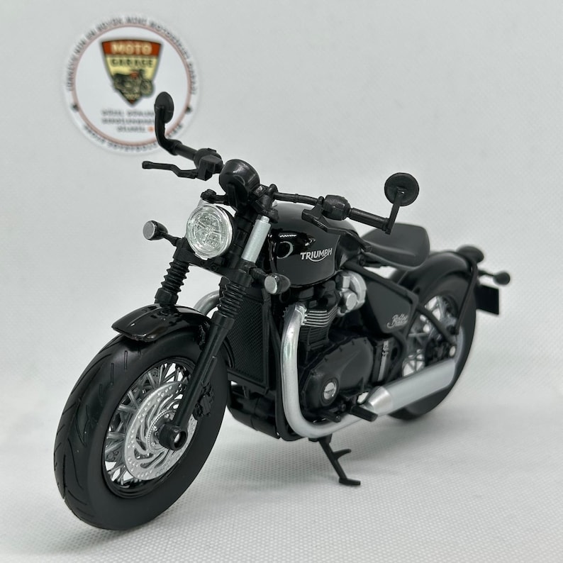 May include: A black Triumph motorcycle model with silver detailing. The model features a round headlight, detailed engine components, and the Triumph logo on the fuel tank. The motorcycle has a classic design with a low seat and a curved exhaust pipe.
