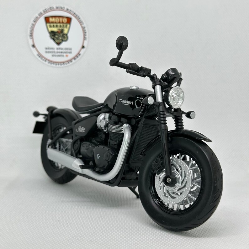 May include: A black Triumph motorcycle model with silver detailing on the exhaust and wheels. The bike features a classic design with a round headlight and a small seat. The word "Triumph" is visible on the fuel tank.