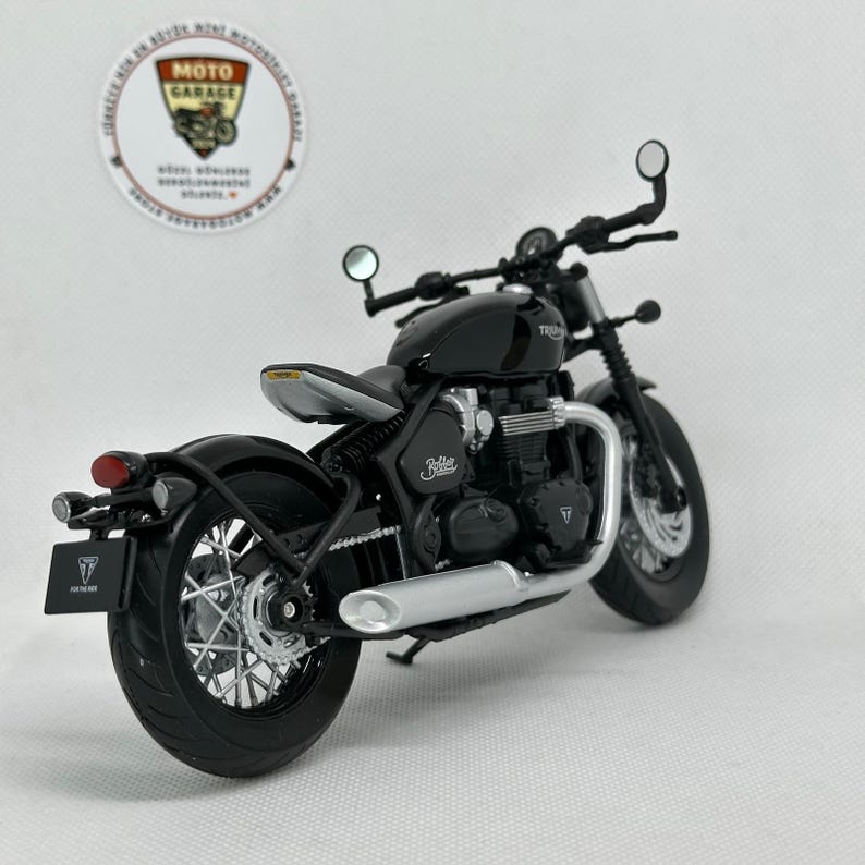 May include: A black motorcycle model with silver detailing, including a round headlight and a curved exhaust. The model has the word "Bobber" on the side and "FOR THE RIDE" on the license plate. The model is a miniature replica.
