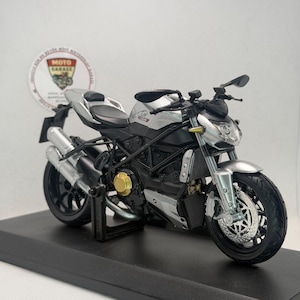 Peut inclure: Maquette de moto argentée et noire avec des détails précis, notamment une selle noire, des pots d'échappement et des roues. Le modèle a un composant moteur doré et est présenté sur une base noire. Le logo "MOTO GARAGE" est également visible.