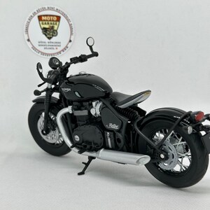May include: A black Triumph Bobber motorcycle model with silver detailing. The model has a detailed engine, exhaust pipe, and wheels. The motorcycle is set against a white background, with a Moto Garage sticker visible.