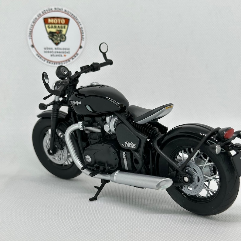 May include: A black Triumph Bobber motorcycle model with silver detailing. The model has a detailed engine, exhaust pipe, and wheels. The motorcycle is set against a white background, with a Moto Garage sticker visible.