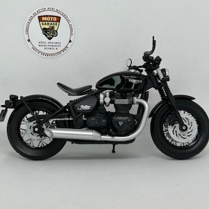 May include: A black Triumph motorcycle model with silver exhaust pipes and a black seat. The model has the word "Triumph" on the gas tank and "Bobber" on the side. A Moto Garage store logo is in the background.