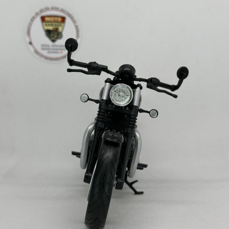May include: A miniature black and silver motorcycle, seen from the front. It has a round headlight, handlebars, and side mirrors. The tyres are black, and the frame is silver. A logo is visible in the background.