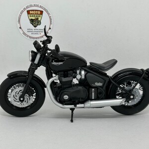 May include: A black Triumph motorcycle model with silver detailing. The model features a detailed engine, wheels, and exhaust pipe. The motorcycle has the word "Triumph" on the gas tank and "Bobber" on the side.
