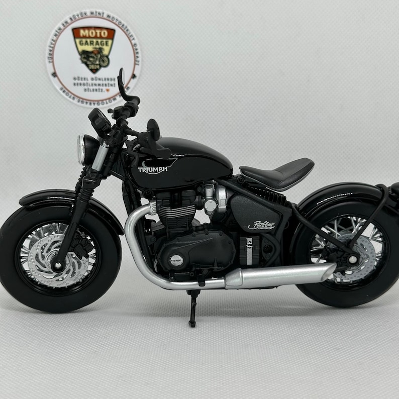 May include: A black Triumph motorcycle model with silver detailing. The model features a detailed engine, wheels, and exhaust pipe. The motorcycle has the word "Triumph" on the gas tank and "Bobber" on the side.