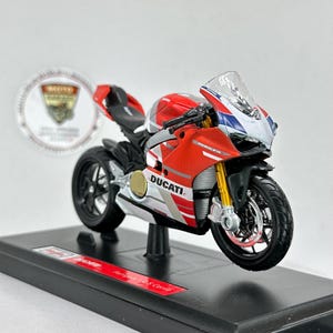 Może przedstawiać: Czerwony, biały i czarny model motocykla Ducati Panigale V4 S Corse. Model posiada przezroczystą szybę przednią, złote przednie widelce i czarne koła. Motocykl jest zamontowany na czarnym stojaku wystawowym.