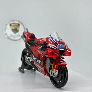 Peut inclure: Modèle réduit de moto Ducati rouge avec des roues noires et un pare-brise transparent. Le modèle présente le logo Lenovo et le numéro 43. La moto est sur un support noir.