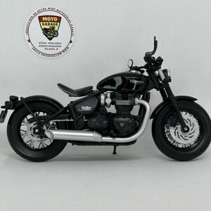 May include: A black Triumph motorcycle model with silver exhaust pipes and a black seat. The model has the word "Triumph" on the gas tank and "Bobber" on the side. A Moto Garage store logo is in the background.