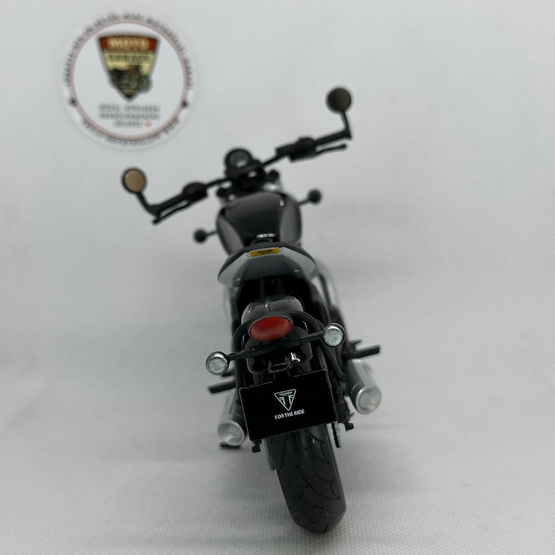May include: A black motorcycle model with a red rear light and silver exhaust pipes. The model has a license plate that reads "FOR THE RIDE" and a logo. The handlebars have round mirrors.