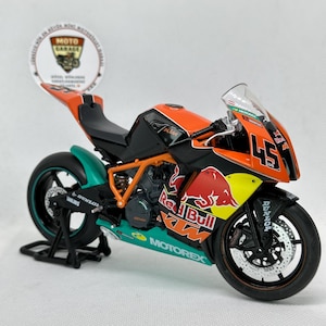 Peut inclure: Modèle réduit de moto aux couleurs orange, noir et turquoise, avec les logos Red Bull et KTM. Le numéro 45 est affiché sur le carénage avant. Le modèle est présenté sur un support noir, mettant en valeur les détails d'une moto de course.