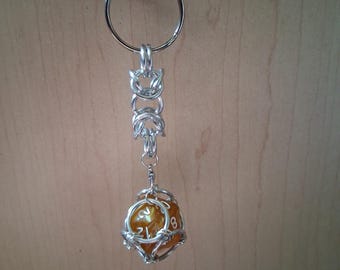 May include: A silver-toned metal keychain featuring a chainmail-style link connector attached to a spherical cage. Inside the cage sits a translucent amber-colored twenty-sided die with white numbers, suspended from a circular metal ring.