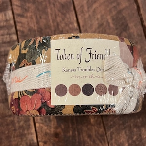 Peut inclure: Un paquet de tissu enveloppé dans du plastique transparent, avec des motifs floraux rouges, verts et dorés. L'étiquette indique "Token of Friend" et "Kansas Troubles Quilts". Le paquet est attaché avec un ruban crème.