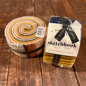 Puede incluir: Dos paquetes de tela sobre una superficie de madera. Uno es un rollo en espiral de tiras de tela coloridas. El otro es un cuaderno de bocetos envuelto en plástico, con el texto "sketchbook" y "Ruby Society" visible.