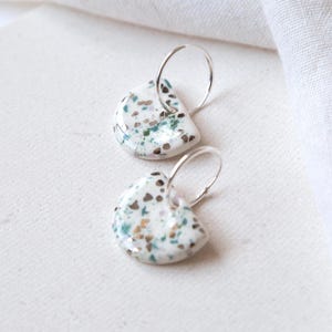 May include: A pair of ceramic earrings with a speckled design. The earrings have a white base with teal, brown, and gold speckles. Each earring is a half-circle shape and hangs from a silver hoop.