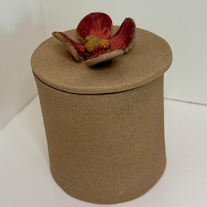 Op de afbeelding: Een kleine, cilindrische, bruinbeige keramische pot met een deksel. Het deksel is bekroond met een rood en geel bloemvormig ornament. De pot staat op een wit oppervlak.