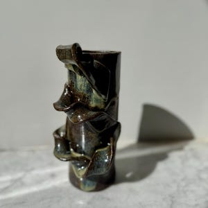 Peut inclure: Un vase en céramique fait à la main, de forme cylindrique et à la finition sombre et brillante. Le vase présente des éléments texturés et pliés autour de son corps, avec des touches de glaçure verte et bleue. Le design est organique et sculptural.