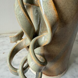 Könnte beinhalten: Eine handgefertigte Keramikvase mit einem einzigartigen, fließenden Design. Die Vase hat eine hellgrüne und braune Farbgebung mit einer strukturierten Oberfläche. Die Griffe werden durch schleifenartige, bandförmige Verlängerungen gebildet, die sich krümmen und verdrehen.