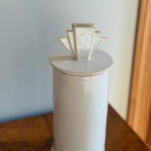 Puede incluir: Un tarro cilíndrico de cerámica blanquecina con tapa. La tapa presenta un diseño geométrico en forma de abanico. El tarro está moteado y se encuentra sobre una superficie de madera. El diseño es minimalista y moderno.