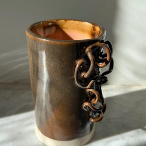 May include: A handmade ceramic mug with a dark brown glaze and a unique, swirling handle. The interior has a pinkish hue, and the base is a creamy white. This artful mug is perfect for coffee or tea.