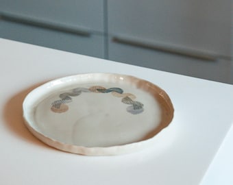 May include: A round, off-white ceramic plate with a slightly ruffled edge. The plate features a decorative pattern of interconnected, semi-circular designs in shades of blue, brown, and gray. The plate is on a white surface.