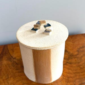 May include: A cylindrical ceramic jar with a matte off-white finish and a vertical brown stripe. The lid features a small, arched handle constructed from several tiny, stacked square tiles in shades of tan, blue, and white.