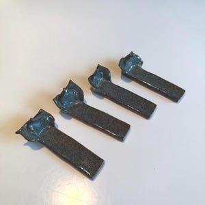 Peut inclure: Quatre repose-baguettes en céramique. Chaque repose-baguettes est de couleur gris foncé avec une base rectangulaire et un dessus évasé en forme de feuille. Les repose-baguettes sont disposés en diagonale sur une surface blanche.
