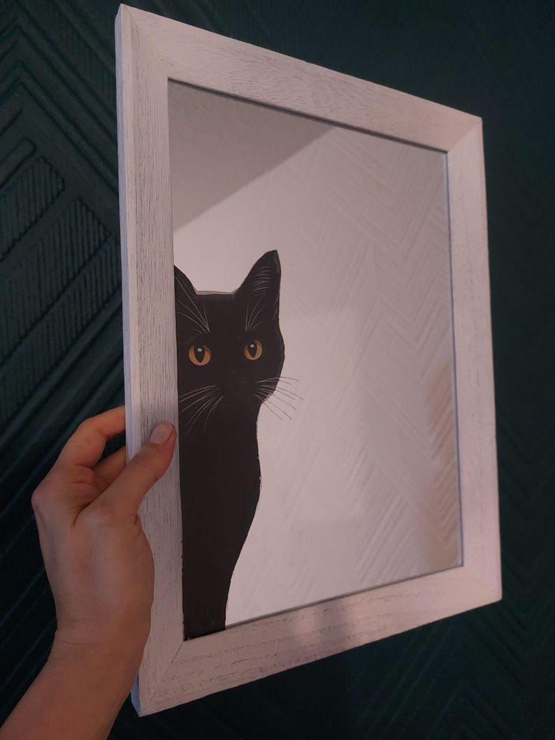 Op de afbeelding: Een ingelijste spiegel met een zwarte kat die van opzij kijkt. De kat heeft gele ogen en witte snorharen. De lijst is wit en de achtergrond van de spiegel is een wit patroon.