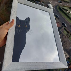 Op de afbeelding: Een rechthoekige spiegel met een lichtgrijs houten frame. De spiegel reflecteert een bewolkte lucht en toont een zwart katten silhouet met gele ogen die van de zijkant gluurt. Een decoratief item.