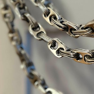 Puede incluir: Un collar de cadena de plata hecho de múltiples bucles de metal entrelazados. La cadena es gruesa y tiene un acabado brillante.