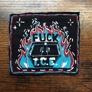 May include: Black square patch with red and blue stitching. The patch features the word "FUCK" above a blue and white image, with the letters "T.C.E." below. Flames and white stars surround the image.