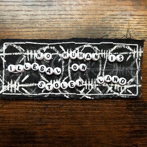 May include: A black patch with white stitching and a white geometric design. The patch features the text "NO HUMANS IS ILLEGAL ON STOLEN LAND" spelled out with white letter beads.