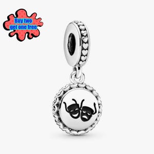 Op de afbeelding: Een zilveren bedel met een theaterthema. De bedel heeft een ronde hanger met twee gestileerde theatermaskers. Het bovenste gedeelte heeft een hartvormig ontwerp. De bedel hangt aan een ring met een hartpatroon.