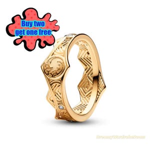 May include: A gold-colored ring with intricate embossed patterns and small clear gemstones. The ring features a repeating geometric design and a decorative emblem. The image includes text that says "Buy two get one free".