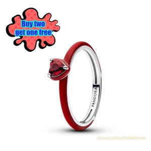 May include: A red and silver ring with a heart-shaped red gemstone. The ring has a red enamel band and the word "PANDORA" is printed on the side. A red and black graphic with the text "Buy two get one free" is in the upper left corner.